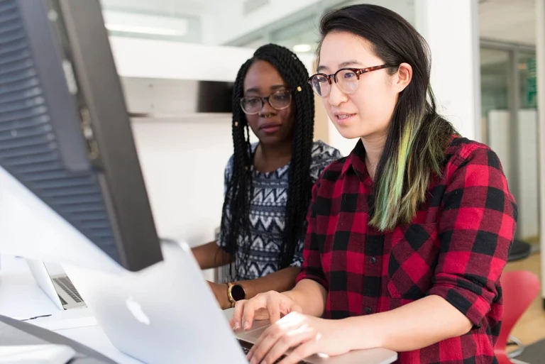 A Black woman and an East Asian woman stand next to each other at a desktop screen, working.