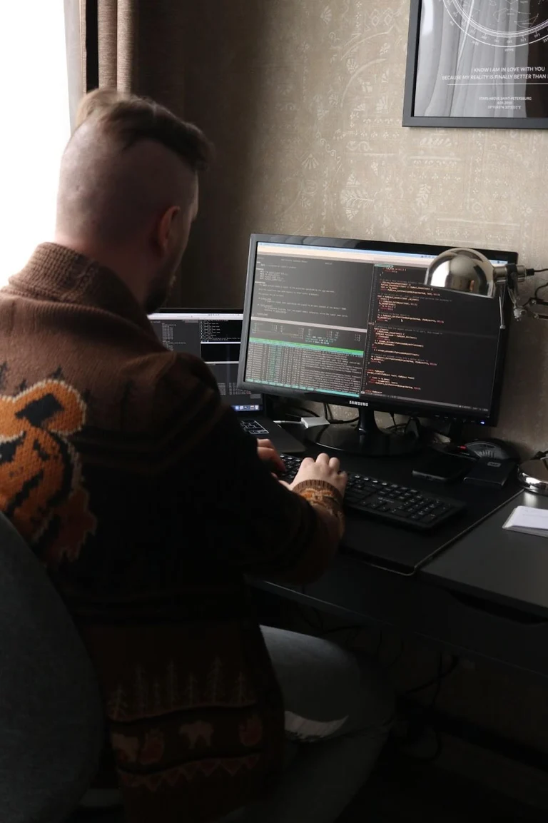 A programmer sits at their desk near a window writing code, back facing the viewer.