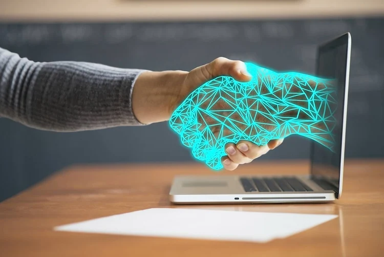 One arm physically shakes hands with a virtual arm coming out of their laptop screen, symbolizing how if you choose to outsource, benefits include the ability to tap into and work alongside talent from anywhere in the world.