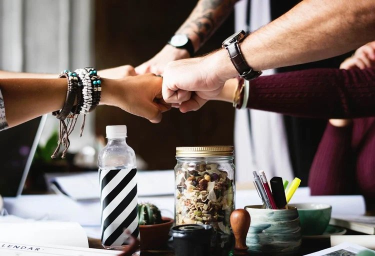 A team of five professionals, whose arms are the only thing visible, join fists at the center of a table to show their willingness to work together and joining forces, demonstrating how when a team decide to outsource, benefits make everyone's work easier.