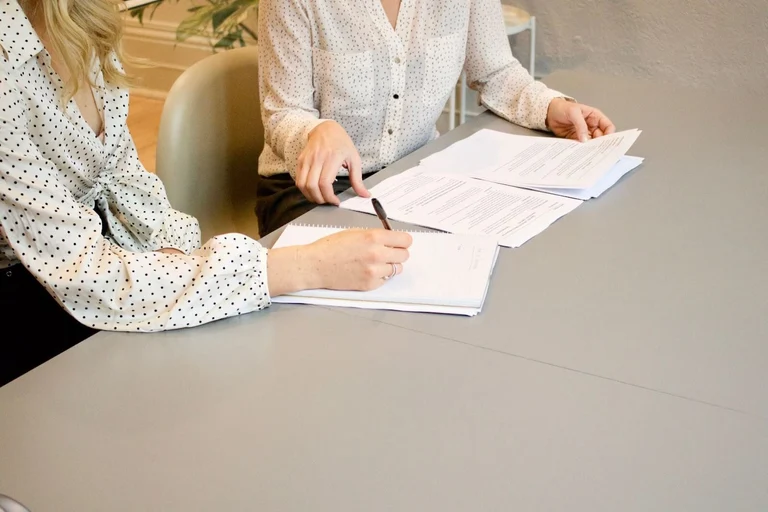 Two women sit side-by-side and go over administrative paperwork together.