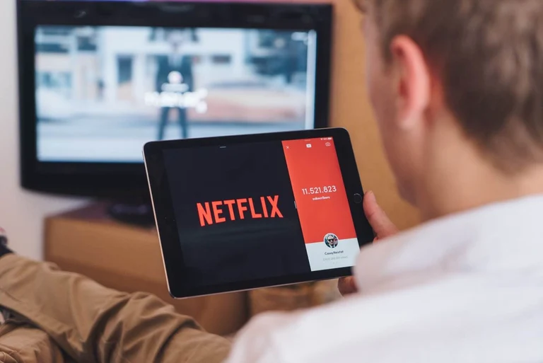A person sits and looks at the Netflix app on his handheld device, with a larger screen in the background. 