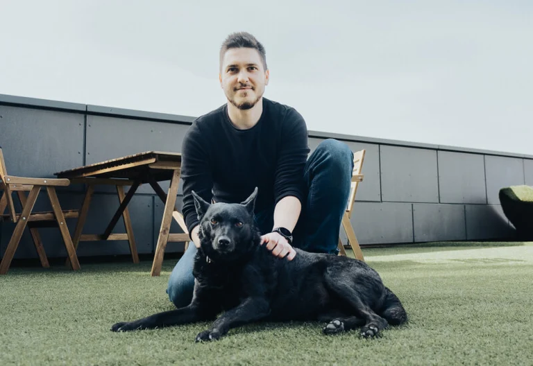 Employee is kneeling in an all-black outfit with his black dog on the grass 