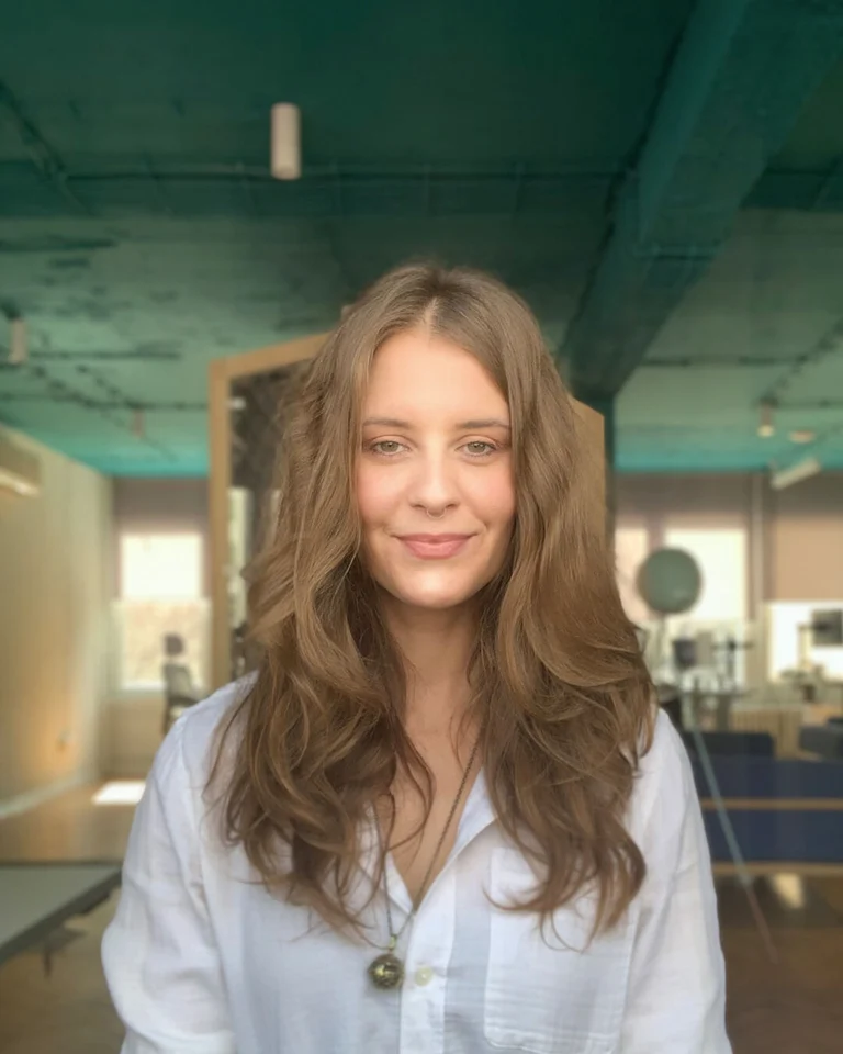 Employee is in a white top looking at the camera with long wavy brown hair