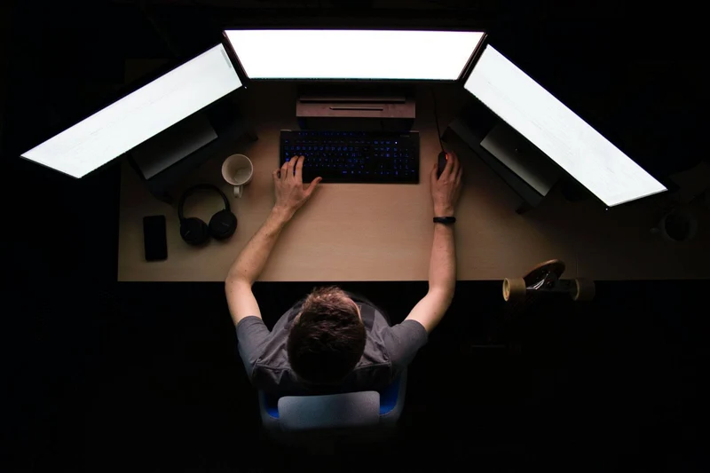 A person sits at three screens in the dark, illuminated by the light produced by the screens, working on a project. 
