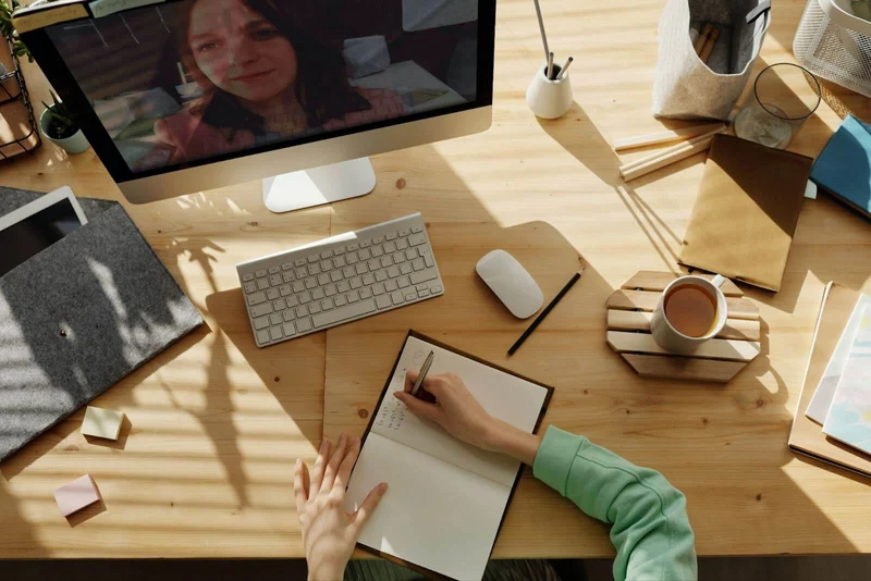A wooden desk shows the work station of a woman of whom only her arms are visible. She's taking notes on a notebook placed at the center of her desk while enjoying a cup of coffee with milk placed to her right, all of this while watching a Zoom call her Mac monitor at the back of the desk.