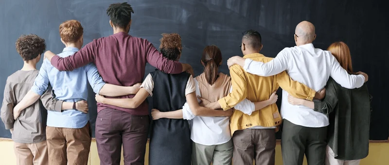 picture of a group of racially diverse and gender diverse individuals standing with their arms together, facing a blank chalkboard