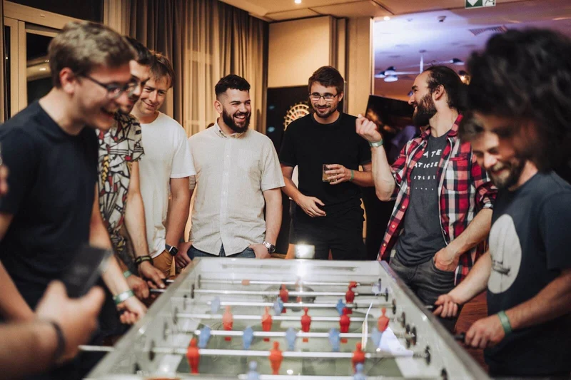 A group of developers stand around a foosball/kicker table in the DO OK office. They are smiling and chatting and two of them go head to head.