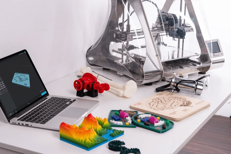 Typical scene found in prototype companies made up of a desk supporting a laptop, a 3D printing machine, and several plastic prototypes.