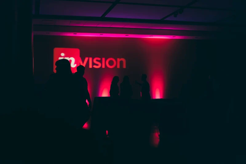 A photograph of a dark room lit by a red neon sign of the InVision logo.