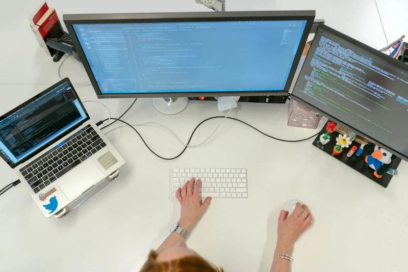 A programmer/developer can be seen writing code. There's a laptop on the left and two monitors that're connected to it, side to side.