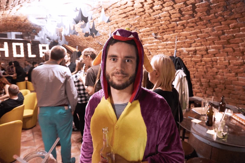 An employee looks directly at the camera in a dinosaur costume with a drink in hand as others are mingling in the background