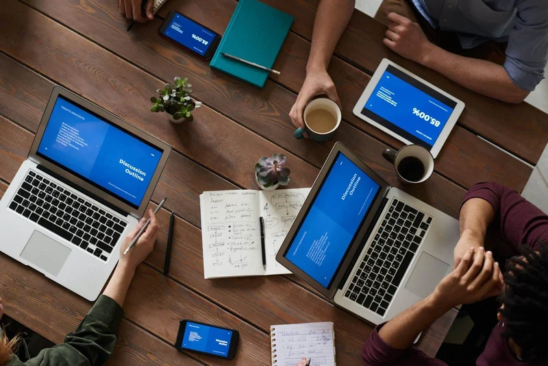 A team of at least 4 people are sitting down in a wooden table that looks like that of a picnic bench. Only their arms are visible. On top of the table, there are pens, notebooks, two cups of coffee, two Macbook laptops, one tablet, and two smartphones, all of which are displaying a blue slideshow. Meetings like these could be replaced by the use Loom and other tools, which would save these coworkers a lot of time.