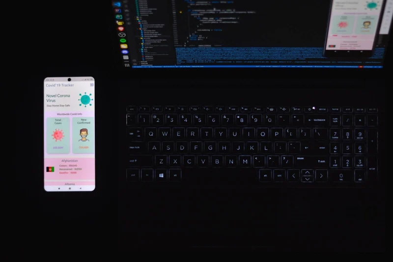 Flutter code on laptop screen with smartphone open on a mobile app next to it. 